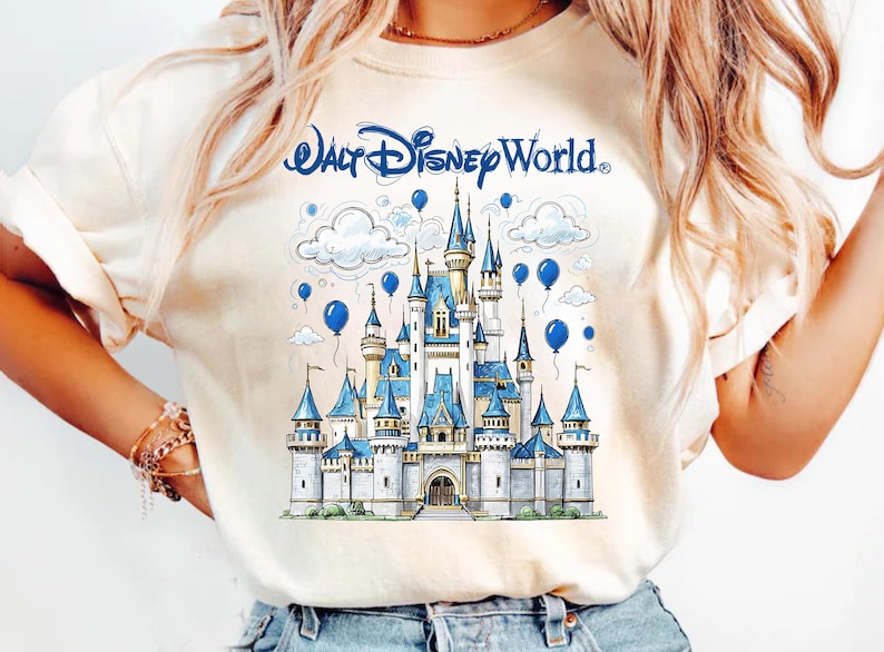 May include: Cream-colored t-shirt featuring a detailed illustration of Cinderella's castle with blue accents, clouds, and balloons. The text "Walt Disney World" is arched above the castle. A whimsical design for Disney fans.