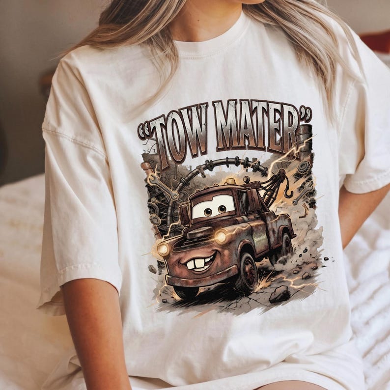 May include: Cream-colored t-shirt featuring a cartoon illustration of Tow Mater from the movie Cars. The graphic includes the text "TOW MATER" in brown lettering, with the character depicted in a dynamic, action-oriented pose with lightning effects.