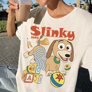 Puede incluir: Camiseta color crema con una ilustraci&oacute;n de dibujos animados de Slinky Dog de Toy Story. El dise&ntilde;o incluye la palabra "Slinky" en naranja, el a&ntilde;o "1995" y elementos tem&aacute;ticos de juguetes como bloques y una pelota.