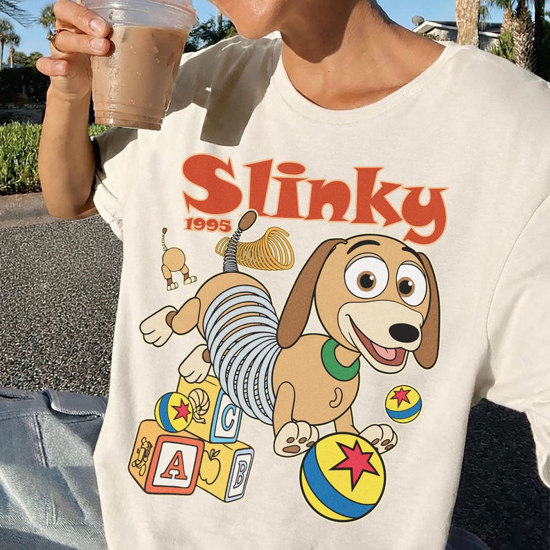 Puede incluir: Camiseta color crema con una ilustraci&oacute;n de dibujos animados de Slinky Dog de Toy Story. El dise&ntilde;o incluye la palabra "Slinky" en naranja, el a&ntilde;o "1995" y elementos tem&aacute;ticos de juguetes como bloques y una pelota.
