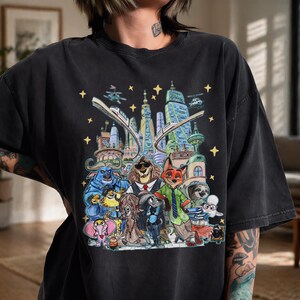 K&ouml;nnte beinhalten: Schwarzes T-Shirt mit einer farbenfrohen Illustration von Zootopia-Charakteren. Das Design zeigt Tierfiguren vor einer Stadtkulisse, komplett mit Sternen und einer Einschienenbahn. Das T-Shirt hat eine lockere Passform.