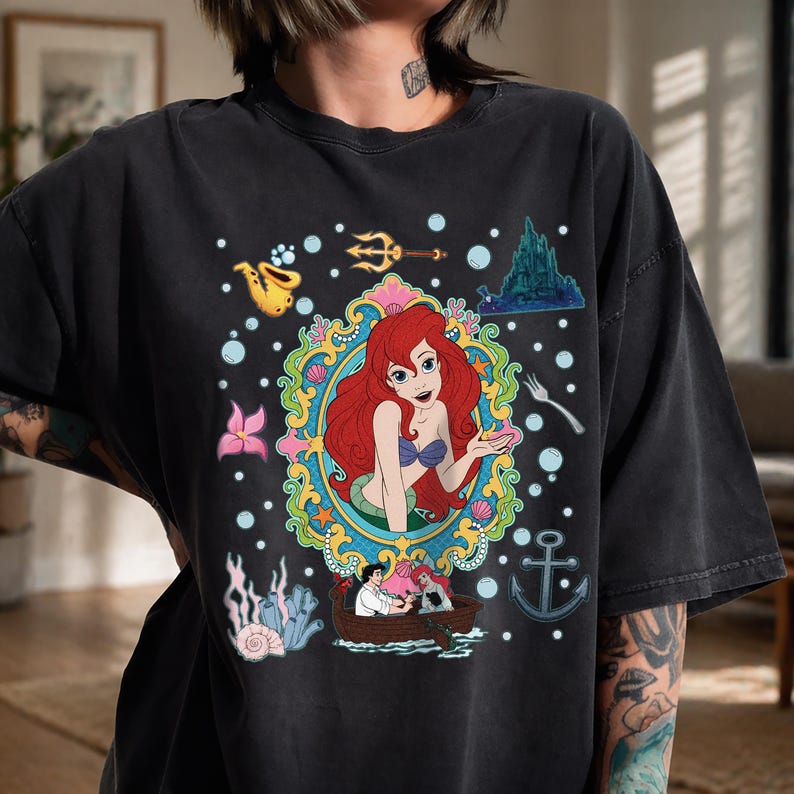 以下が含まれることがあります： 黒いTシャツ。リトルマーメイドのアリエルのカラフルなグラフィックが特徴です。デザインにはアリエル、ボート、城、さまざまな海のテーマの要素が含まれています。グラフィックは泡と装飾的なフレームで囲まれています。