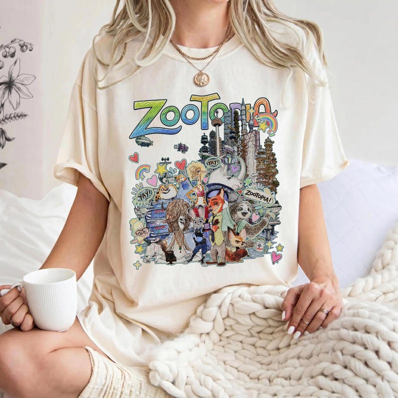 May include: Cream-colored t-shirt featuring a colorful graphic of the word "Zootopia" and various characters from the animated film. The design includes cityscapes, rainbows, and cartoon animals. The shirt is a relaxed fit.