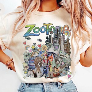 May include: Ivory-colored t-shirt featuring a colorful graphic from the animated film "Zootopia." The design includes the word "Zootopia" in a rainbow gradient, along with various characters and cityscapes from the movie.