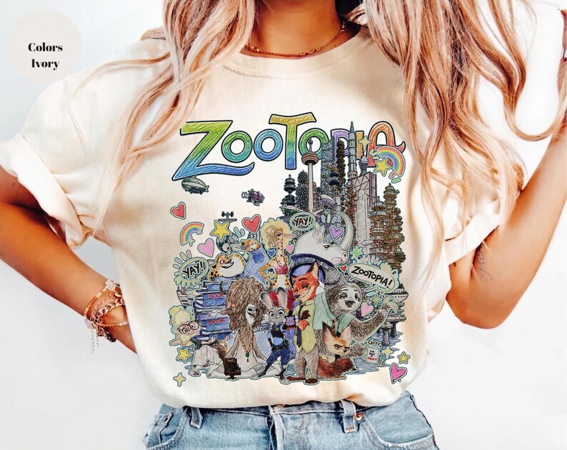 May include: Ivory-colored t-shirt featuring a colorful graphic from the animated film "Zootopia." The design includes the word "Zootopia" in a rainbow gradient, along with various characters and cityscapes from the movie.