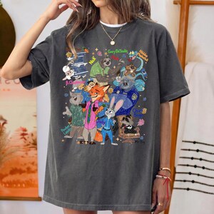 Op de afbeelding: Donkergrijs T-shirt met een kleurrijke illustratie van personages uit de film Zootopia. Het ontwerp bevat Judy Hopps, Nick Wilde en andere personages met hun namen. Het shirt heeft een ronde hals en korte mouwen.