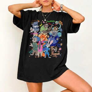 Op de afbeelding: Zwart T-shirt met een kleurrijke cartoonillustratie van personages uit de film Zootopia. De afbeelding bevat verschillende dierenfiguren met hun namen, zoals Nick, Judy en Flash. Het shirt heeft een casual, oversized stijl.