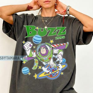 May include: Dark gray t-shirt featuring a graphic of Buzz Lightyear from Toy Story. The design includes the word "Buzz" in green, the year "1995", and images of planets and stars. The shirt has short sleeves.