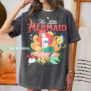 May include: Dark grey t-shirt with a colourful graphic of Ariel from The Little Mermaid. The design includes Ariel, Flounder, Sebastian, and the text "The Little Mermaid" in red and white. The shirt has a relaxed fit.