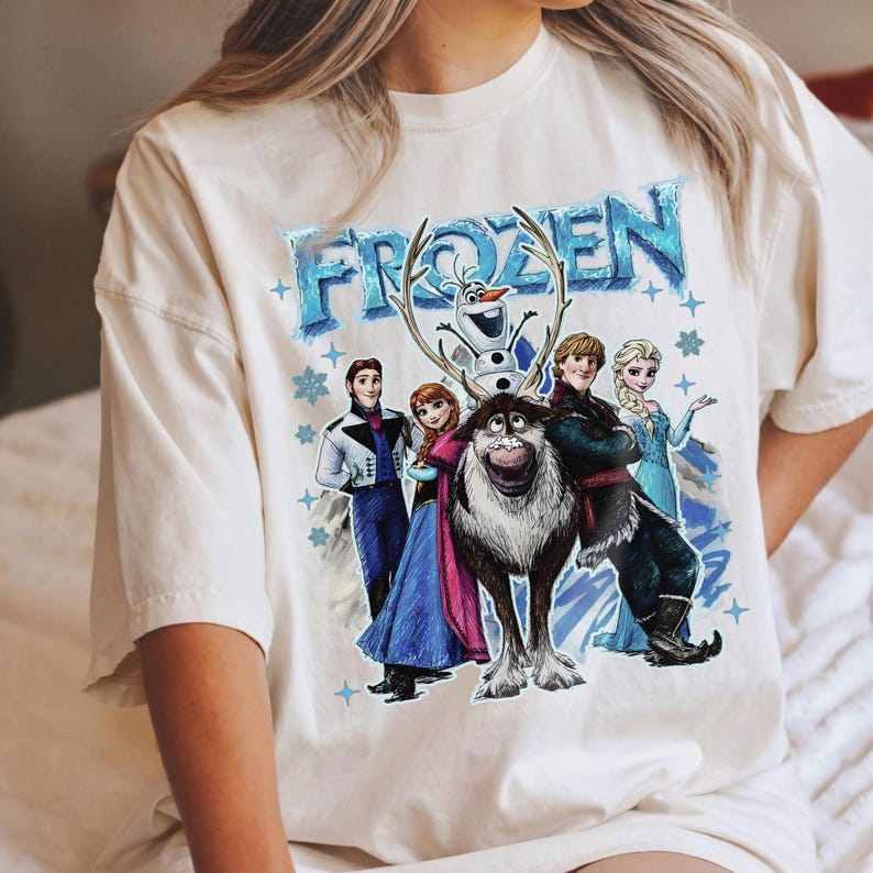 May include: Cream-coloured t-shirt featuring a graphic of characters from the animated film "Frozen." The design includes the word "FROZEN" in blue, along with images of Anna, Elsa, Kristoff, Olaf, and Sven. Blue stars and snowflakes surround the characters.