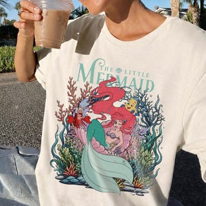 May include: Cream-coloured t-shirt with a vibrant graphic of Ariel from "The Little Mermaid." The design showcases Ariel with red hair, a teal tail, and characters like Sebastian and Flounder, set amongst coral and seaweed.