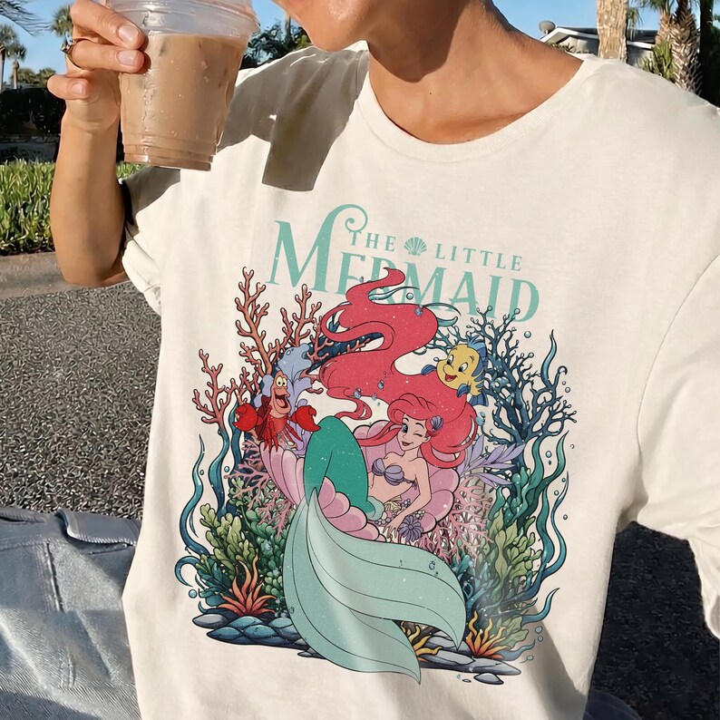 May include: Cream-coloured t-shirt with a vibrant graphic of Ariel from "The Little Mermaid." The design showcases Ariel with red hair, a teal tail, and characters like Sebastian and Flounder, set amongst coral and seaweed.