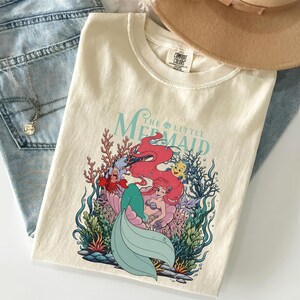 May include: Cream-coloured t-shirt featuring a graphic of Ariel from The Little Mermaid. The design includes Ariel, Sebastian, Flounder, and coral. The words "The Little Mermaid" are in a teal font.
