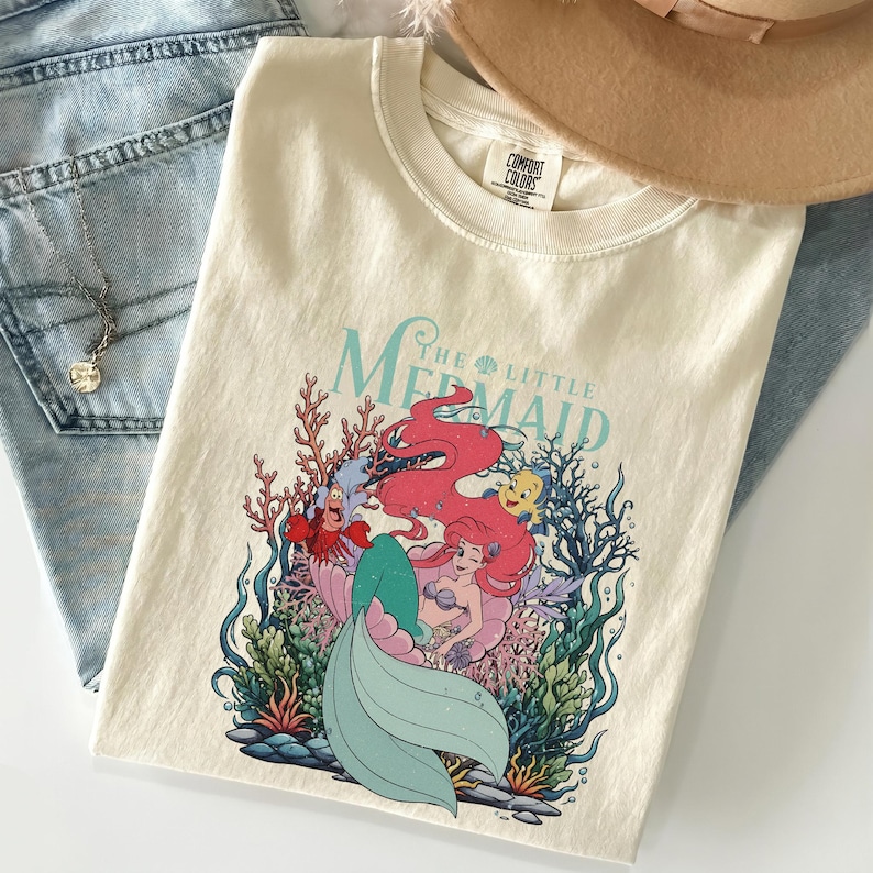 May include: Cream-coloured t-shirt featuring a graphic of Ariel from The Little Mermaid. The design includes Ariel, Sebastian, Flounder, and coral. The words "The Little Mermaid" are in a teal font.