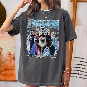 May include: Dark grey t-shirt with the word "FROZEN" in blue, and a graphic of characters from the film. The design includes Elsa, Anna, Kristoff, Sven, and Olaf. The shirt has short sleeves and a relaxed fit.