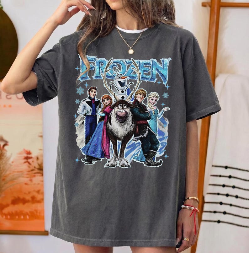 May include: Dark grey t-shirt with the word "FROZEN" in blue, and a graphic of characters from the film. The design includes Elsa, Anna, Kristoff, Sven, and Olaf. The shirt has short sleeves and a relaxed fit.