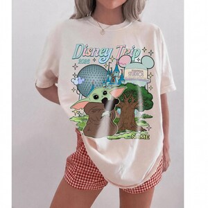 Puede incluir: Camiseta color crema con un gr&aacute;fico de Baby Yoda y los lugares emblem&aacute;ticos de Disney World. El dise&ntilde;o incluye el texto "Disney Trip 2023" en una fuente pastel, junto con la opci&oacute;n de agregar un nombre. La camiseta se combina con pantalones cortos a cuadros rojos y blancos.