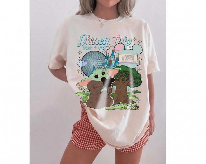 Puede incluir: Camiseta color crema con un gr&aacute;fico de Baby Yoda y los lugares emblem&aacute;ticos de Disney World. El dise&ntilde;o incluye el texto "Disney Trip 2023" en una fuente pastel, junto con la opci&oacute;n de agregar un nombre. La camiseta se combina con pantalones cortos a cuadros rojos y blancos.