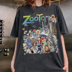 May include: Dark gray t-shirt featuring a colorful graphic from the animated film "Zootopia." The design includes the word "Zootopia" in rainbow colors, along with various characters and cityscapes. The shirt has short sleeves and a relaxed fit.
