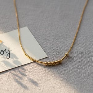 Peut inclure: Un collier doré avec une chaîne délicate et de petits accents perlés. Une carte blanche avec le mot "Joy" en cursive est à côté du collier, ornée d'un petit bouquet de fleurs blanches et d'un ruban beige.