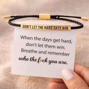 Puede incluir: Pulsera trenzada negra con una barra rectangular dorada grabada con las palabras "DON'T LET THE HARD DAYS WIN". Una tarjeta con texto inspirador se sostiene cerca de la pulsera.