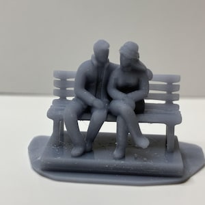 May include: A gray miniature sculpture of a couple sitting on a park bench. The figures are detailed, with the man wearing a jacket and the woman wearing a hat. The bench has horizontal slats and is set on a base.