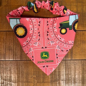 May include: Pink dog bandana with a John Deere logo and tractor design. The bandana features a pink paisley pattern with white floral accents and green and yellow tractor graphics. The elastic band is pink with a green, yellow, and black pattern.