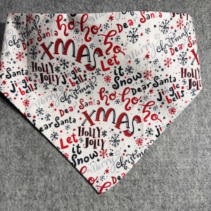 May include: A triangular pet bandana featuring a white background with a repeating Christmas-themed pattern in red, black, and gray. The print includes snowflakes and text such as XMAS, Ho Ho, Dear Santa, Holly, Jolly, Jingle bells, and Let it Snow.