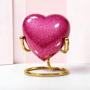 May include: A pink glitter heart-shaped decorative object on a gold-colored stand. The heart has a glossy finish and is supported by a curved, gold-toned metal structure. The base is a gold ring.