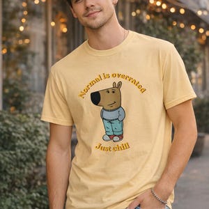 Puede incluir: Camiseta de color amarillo mostaza con una ilustración de dibujos animados de un animal marrón con un suéter azul y pantalones turquesa. El texto "Normal is overrated" y "Just chill" están impresos en la camiseta. La persona lleva vaqueros azules.