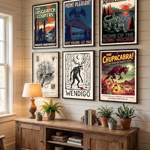 Może przedstawiać: Sześć oprawionych plakatów z ilustracjami kryptyd: Visit Sasquatch Country, Point Pleasant West Virginia, Rhinelander Home of the Hodag, kałamarnica olbrzymia, Wendigo oraz The Chupacabra, zawieszonych na białej ścianie nad drewnianą konsolą.