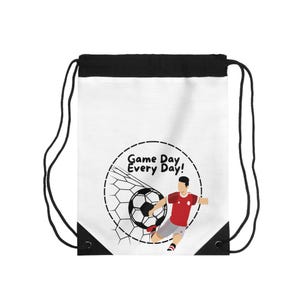 Könnte beinhalten: Weißer Kordelzugrucksack mit schwarzem Oberteil und Kordelzügen. Die Tasche zeigt eine Fußballgrafik mit einem Spieler, der einen Ball in ein Netz schießt. Der Text lautet "Game Day Every Day!"