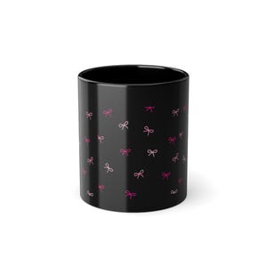 Könnte beinhalten: Schwarze Keramiktasse mit einem sich wiederholenden Muster aus rosa Schleifen. Die Schleifen sind in verschiedenen Rosatönen gehalten und gleichmäßig über die Oberfläche der Tasse verteilt. Die Tasse hat eine glänzende Oberfläche und eine einfache, zylindrische Form.