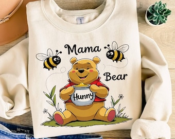 May include: A cream-coloured crewneck sweatshirt featuring a graphic of a yellow bear wearing a red shirt, holding a white honey pot labelled Hunny. The design includes two bees, green grass, and the text Mama Bear in black cursive lettering.