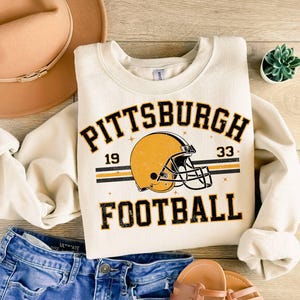 May include: A cream-colored crewneck sweatshirt featuring a graphic of a yellow football helmet with black accents. The text reads PITTSBURGH 19 33 FOOTBALL in black block lettering. The design includes thin horizontal stripes and small star icons.
