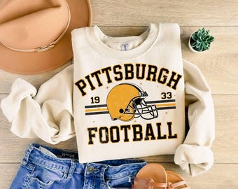 May include: A cream-coloured crewneck sweatshirt featuring a graphic of a yellow football helmet with black accents. The text reads PITTSBURGH 19 33 FOOTBALL in black block lettering. The design includes thin horizontal stripes and small star icons.