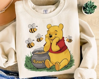 May include: A cream-coloured crewneck sweatshirt featuring a graphic of Winnie the Pooh sitting in green grass next to a grey honey pot labelled HUNNY. Three small yellow and black bees fly around the character, who wears a red shirt.