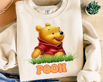 May include: A cream-coloured crewneck sweatshirt featuring a graphic of the yellow bear character Winnie the Pooh wearing a red shirt, positioned above green grass and the orange text POOH in a retro font, with a tag visible at the neckline.