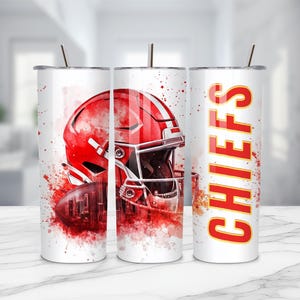 May include: A white cylindrical tumbler with a metal straw featuring a red football helmet graphic, a football, and red watercolor splatter accents. The word CHIEFS is printed vertically in bold red letters with a yellow outline on the side.