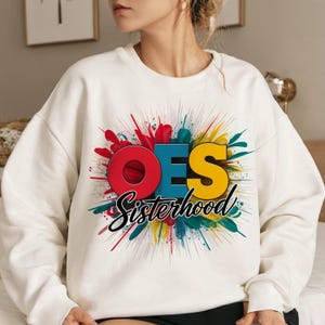 May include: A cream-colored crewneck sweatshirt featuring a graphic design with the letters OES in red, blue, and yellow. Below the letters, the word Sisterhood is written in black script. The design includes a colorful paint splatter background.