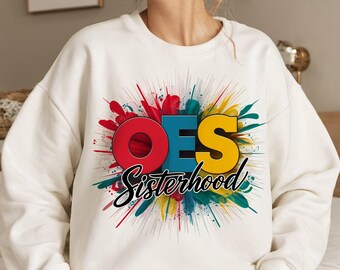 May include: A cream-coloured crewneck sweatshirt featuring a graphic design with the letters OES in red, blue, and yellow. Below the letters, the word Sisterhood is written in black script. The design includes a colourful paint splatter background.