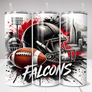 May include: A cylindrical tumbler with a straw featuring a graphic print of a black football helmet, a brown football, a stadium light, and a city skyline. The design includes red and black paint splatters and the word FALCONS in white text.