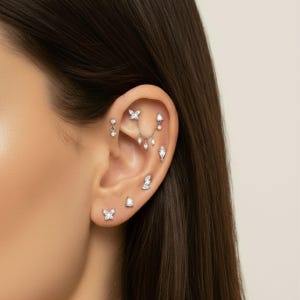 May include: Close-up of an ear adorned with multiple pieces of jewelry. The ear features several small, sparkling earrings, including butterfly and teardrop designs. The jewelry is silver-toned and set with clear gemstones, creating a delicate and elegant look.