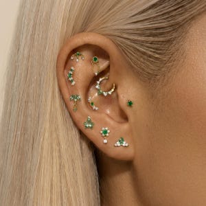 May include: An ear displaying various earrings. The jewellery incorporates emerald green gemstones and small white stones, set in gold. Designs include studs, hoops, and dangling styles, creating a layered and elegant appearance.