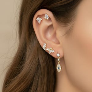 May include: Close-up of a person's ear adorned with multiple gold earrings. The earrings feature clear, sparkling gemstones in various shapes, including teardrops, triangles, and floral designs. One earring has a blue center. The jewelry is delicate and elegant.