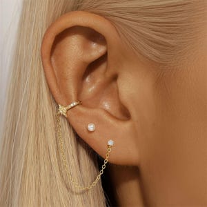 May include: A close-up of an ear adorned with gold jewelry. A star-shaped ear cuff with a chain connects to a small square stud earring. A pearl stud earring is also visible. The jewelry is set against a background of blonde hair.