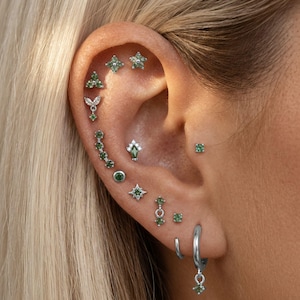 Può includere: Primo piano di un orecchio adornato con molteplici gioielli. L'orecchio presenta vari orecchini a lobo, cerchi e pendenti, tutti in argento con pietre preziose verdi. I gioielli includono stelle, fiori e forme geometriche.