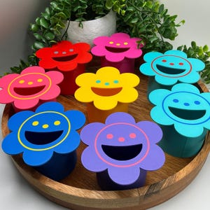 May include: A collection of seven cylindrical containers with flower-shaped lids in red, pink, yellow, purple, blue, and two shades of teal. Each lid features a smiling face with a wide mouth, two eyes, and a nose, rendered in contrasting colours.