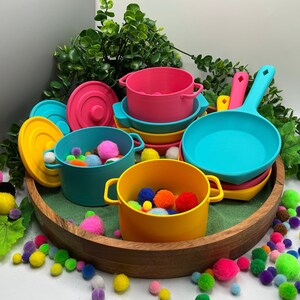 May include: A collection of colourful plastic toy cookware including pots with lids and a frying pan in shades of pink, yellow, and turquoise. The containers are filled with small, multi-coloured fuzzy pom-poms, arranged in a wooden bowl.