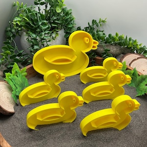 May include: A selection of bright yellow duck-shaped cookie cutters. The set includes a large duck outline and several smaller duck shapes. The cutters are made of plastic and are arranged on a dark surface with greenery in the background.